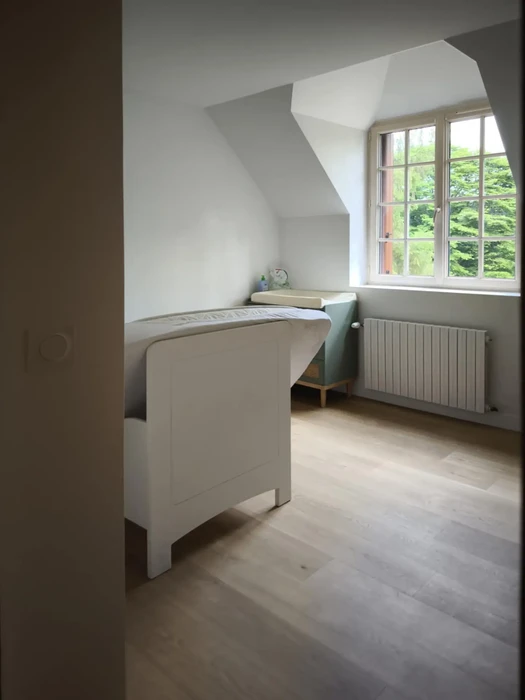 Salle de bain moderne sous combles avec baignoire îlot blanche, radiateur horizontal blanc, fenêtre à croisillons donnant sur jardin arboré
